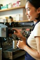 Lycklig asiatisk kvinna, Barista använder sig av kaffe maskin till göra ordning, ångande mjölk för cappuccino och latte, skrattande och leende medan arbetssätt i Kafé foto