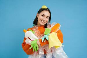 porträtt av leende asiatisk kvinna kramas tömma plast flaskor och återvinningsbar sopor, ekoaktivist gillar till återvinna, blå bakgrund foto
