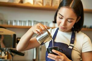 leende asiatisk Barista flicka, framställning kaffe, häller ångad mjölk in i cappuccino, håller på med latte konst i kopp, arbetssätt i Kafé foto