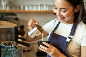 leende asiatisk Barista flicka, framställning kaffe, häller ångad mjölk in i cappuccino, håller på med latte konst i kopp, arbetssätt i Kafé foto