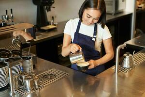 leende asiatisk flicka Barista, Kafé personal häller ångad mjölk i kaffe, förbereda cappuccino med latte konst, stående i blå förkläde Bakom disken foto