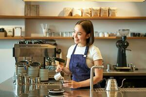 Lycklig leende Kafé ägare, flicka Barista i förkläde, framställning cappuccino, latte konst med ångad mjölk, stående Bakom disken foto