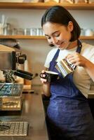 stänga upp av söt asiatisk Barista flicka framställning cappuccino, håller på med latte konst i kopp med ångad mjölk, stående i kaffe affär Bakom disken foto