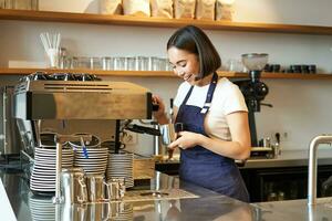 Lycklig asiatisk flicka Barista, arbetssätt i Kafé, bryggning kaffe, förbereda cappuccino, använder sig av ångkokare på maskin till förbereda latte, leende och skrattande foto