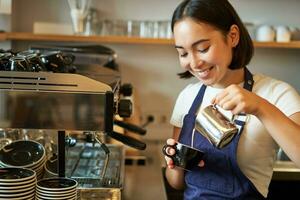 stänga upp av söt asiatisk Barista flicka framställning cappuccino, håller på med latte konst i kopp med ångad mjölk, stående i kaffe affär Bakom disken foto