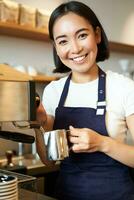 vertikal skott av Barista, asiatisk flicka ångande mjölk för cappuccino, förbereda latte för klient, bär blå förkläde, leende lyckligt, arbetssätt i Kafé foto