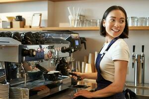 leende asiatisk Barista flicka gör cappuccino med kaffe maskin, står Bakom disken i Kafé foto