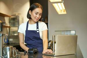 leende asiatisk flicka Barista, arbetssätt i Kafé, tjänande klient, ser på bärbar dator medan använder sig av kort läsare till motta pengar för ordning, försäljning kaffe foto