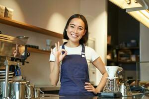 Barista på de disken visar Okej tecken. leende asiatisk flicka Arbetar i Kafé, bär blå förkläde, rekommenderar henne kaffe affär foto