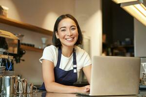 entusiastisk asiatisk flicka i Kafé enhetlig, Barista arbetstagare med bärbar dator, ser Lycklig och överraskad på kamera foto