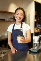leende asiatisk flicka Barista ger beställa till klient, innehav hämtmat kaffe kopp, bär förkläde, arbetssätt Bakom disken i Kafé foto