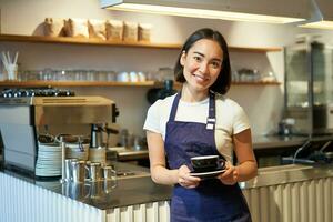 porträtt av asiatisk leende kvinna Barista, bär Kafé förkläde och innehav kopp av kaffe, tar klient beställa till de tabell, tjänande drycker foto