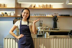 porträtt av asiatisk flicka i förkläde, stående i Kafé nära disken, Barista pekande finger på kopia Plats, tömma baner kaffe affär foto