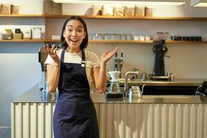 entusiastisk ung asiatisk kvinna, Barista i förkläde, ser förvånad, klappa händer och leende, stående i kaffe affär, arbetssätt i Kafé foto