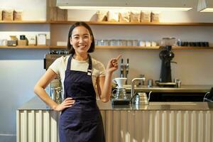 porträtt av asiatisk flicka i förkläde, stående i Kafé nära disken, Barista pekande finger på kopia Plats, tömma baner kaffe affär foto