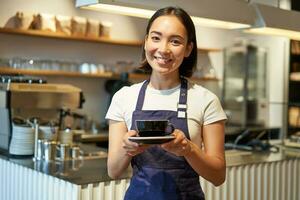 leende asiatisk flicka Barista framställning kaffe, innehav kopp, bär Kafé enhetlig förkläde, stående nära disken foto