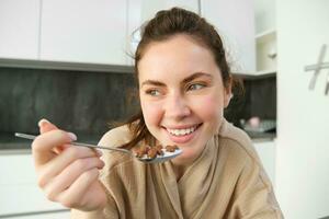 bild av Lycklig ung kvinna äter spannmål på Hem med mjölk, har henne frukost, bär morgonrock, Sammanträde i kök ensam foto