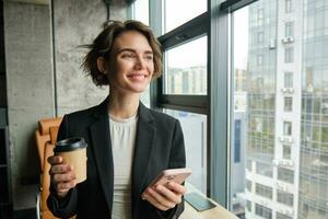 porträtt av framgångsrik kvinna entreprenör drycker kopp av kaffe i ett kontor, innehar smartphone, väntar för klient i konferens rum foto