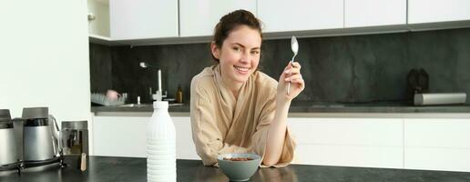 porträtt av skön ung och friska kvinna i morgonrock äter henne frukost i kök, har spannmål med mjölk och leende foto