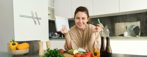 leende ung kvinna i morgonrock, matlagning i de kök, stående nära grönsaker med anteckningsbok, skrivning ner recept, kontroll matvaror lista, matlagning färsk och friska måltid foto