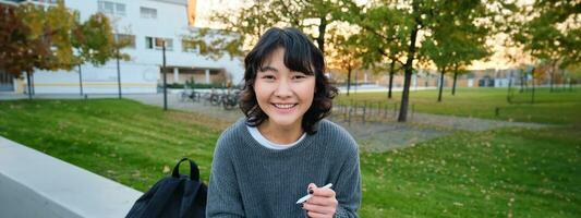 ung asiatisk flicka med grafisk penna och läsplatta, sitter i parkera på bänk, drar repor, gör henne läxa utomhus foto
