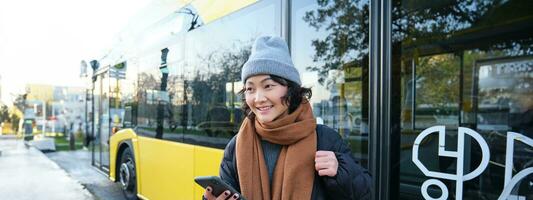 porträtt av flicka stående nära buss på en sluta, väntar för henne offentlig transport, schecks schema på smartphone Ansökan, innehar mobil telefon, bär värma kläder foto
