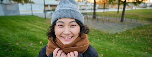 porträtt av söt asiatisk flicka i hatt och scarf, promenader runt om stad i kyligt vår väder, ler och utseende Lycklig, sitter i parkera nära grön gräs foto