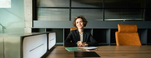 porträtt av affärskvinna i kostym, arbetssätt på dokument i ett kontor, leende på kamera, ser självsäker foto