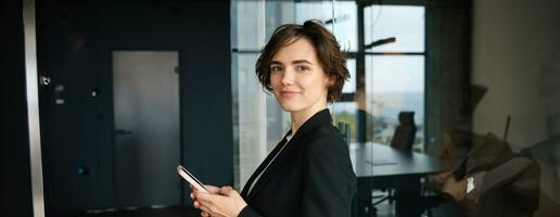 bild av företags- kvinna, försäljare i företag företag, stående i svart kostym, innehav smartphone, kallelse klient foto