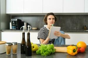porträtt av tänkande kvinna med anteckningsbok, matlagning, skrivning ner recept Ingredienser, beslutar på en måltid för middag, Sammanträde nära grönsaker och hackning styrelse i kök foto