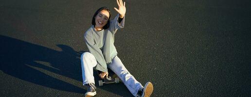 positiv koreanska flicka omslag henne ansikte från solljus, sitter på skateboard och ler lyckligt foto