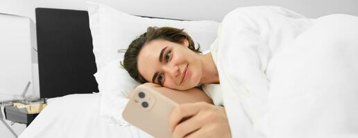 bild av kvinna modell i säng, ser på mobil telefon, läsning på smartphone app, leende nöjd foto