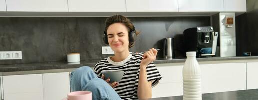 skön ung kvinna modell, äter frukost i de kök, har spannmål med mjölk, leende och dricka kaffe, lyssnande musik eller podcast i trådlös hörlurar foto