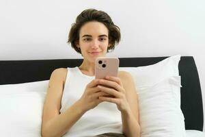 porträtt av leende brunett kvinna vilar i henne sovrum, använder sig av mobil telefon och liggande i säng i henne Hem utrusta, meddelanden, sändning meddelande på smartphone app foto