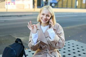 Lycklig skön ung kvinna Sammanträde på bänk med mobil telefon, talande på mobiltelefon och som visar Okej gest foto