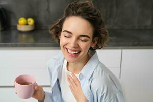 Lycklig morgnar. porträtt av Lycklig brunett kvinna, drycker kopp av kaffe i henne kök och leende, mysigt och värma Start av de dag med cuppa foto