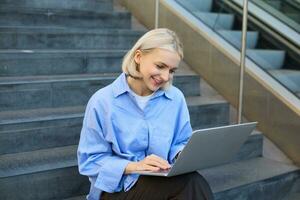 leende flicka studerande använder sig av bärbar dator dator modern teknologi enhet utomhus- i universitet campus uppkopplad inlärning, elearning utanför Sammanträde på urban trappa. webb utbildning kurs webinarier begrepp foto