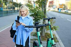 bild av leende ung kvinna rubrik till arbete, hyra, använder sig av mobil telefon app till låsa upp gata skoter till skaffa sig till henne destination, Framställ på gata med ryggsäck foto