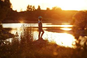 flicka i hatt står på flodstranden vid solnedgången foto