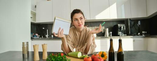 porträtt av kvinna kontroll matvaror lista, ser på grönsaker, innehav anteckningsbok, läsning recept medan matlagning måltid i de kök, hackning tomater och zucchini foto