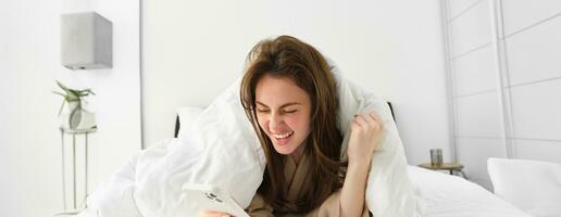 kvinna med smartphone, känsla spänning, läsning mobil telefon och glädjande, liggande i säng under vit filt, använder sig av smartphone och ser Lycklig i morgon- foto