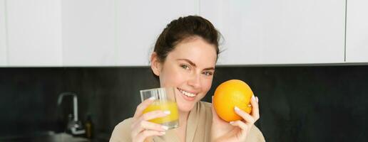 bild av snygg friska kvinna i morgonrock, dricka färsk juice, som visar orange frukt, Framställ i kök foto