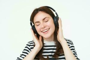 porträtt av skön kvinna i trådlös hörlurar, lyssnande musik, använder sig av hörlurar, leende på kamera, stående över vit bakgrund foto