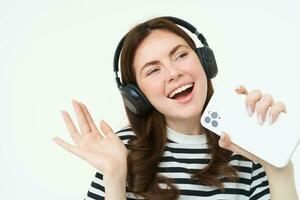 teknologi och social media. porträtt av Lycklig ung kvinna sång karaoke, spelar musik app på smartphone, bär trådlös hörlurar, stående över vit bakgrund foto