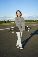 vertikal skott av skater flicka Framställ med longboard, cruising på tömma väg i förorter. leende asiatisk kvinna skridskoåkning på skateboard, innehav kryssare i händer foto