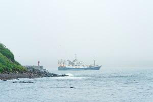 fiske fartyg framträder från Bakom en cape med en fyr, segling in i en dimmig hav foto