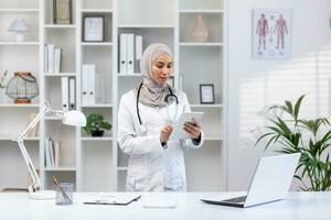 muslim kvinna i vit medicinsk täcka stående inuti medicinsk kontor av klinik, kvinna läkare använder sig av läsplatta dator tänkande och koncentrera sig, koncentrerad kvinna på arbete i sjukhus foto