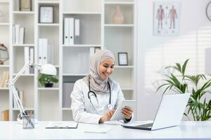 porträtt muslim kvinna läkare i hijab, kvinna läkare med läsplatta dator använder sig av Ansökan, leende nöjd arbete arbetssätt inuti medicinsk kontor, i vit medicinsk täcka, Sammanträde på tabell med bärbar dator foto