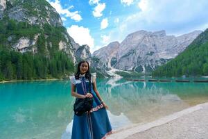 Lycklig asiatisk kvinna turist stående på de kust av braies sjö. dolomiterna, Italien. landskap med flicka, känd sjö med skön reflexion i vatten, träd, himmel med moln. resa. foto