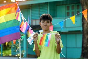 asiatisk pojke innehar en regnbåge flagga i främre av en hus dekorerad med regnbåge flaggor under stolthet månad till visa HBTQ stolthet och identitet. mjuk och selektiv fokus. foto